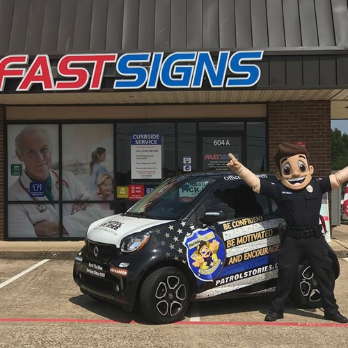 Patrol Stories mascot standing in front of wrapped vehicle at FASTSIGNS of Allen, TX